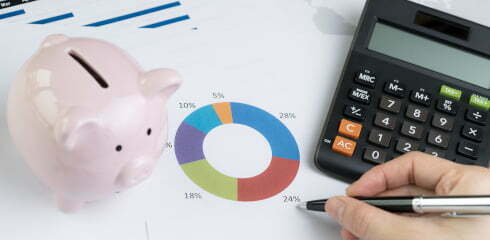 Finance, money budget planning or investment asset allocation concept, hand holding pen reviewing pie chart and graph with calculator and pink piggy bank on table